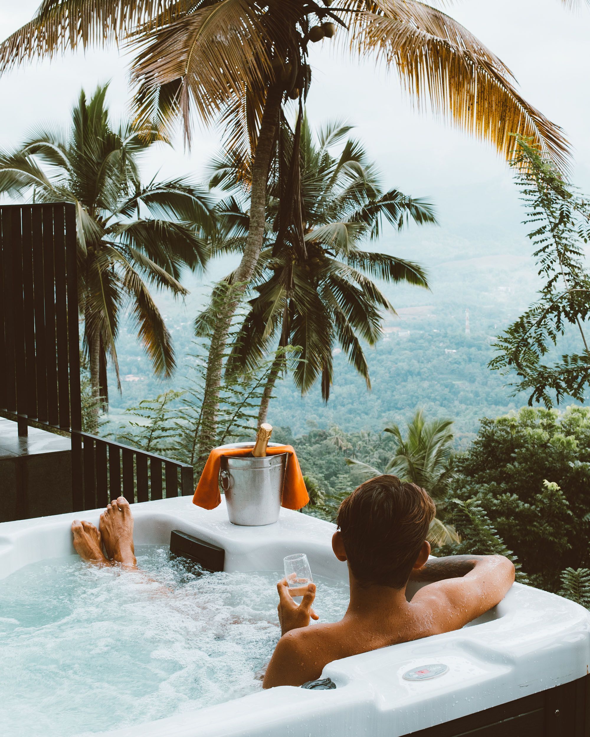 A person relaxing in a hot tub with a view of palm trees and a mountainous landscape.