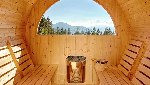 Una sauna de madera con bancos y una piedra central para calentar, con montañas y árboles de fondo.