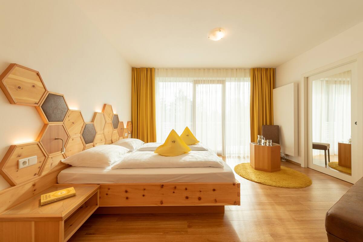 Una habitación de hotel moderna con una cama de madera, una mesita de noche con un libro, una mesita auxiliar de madera, un tapete amarillo y una ventana grande con cortinas.