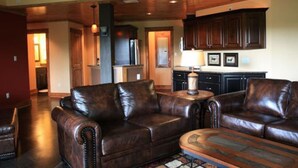 A spacious living room with leather sofas and a coffee table, wooden flooring, and a kitchen area with dark cabinets and a countertop.