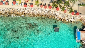 Luchtbeeld van een strand met helder turquoise water, een stenen muur en een rij rode parasols.