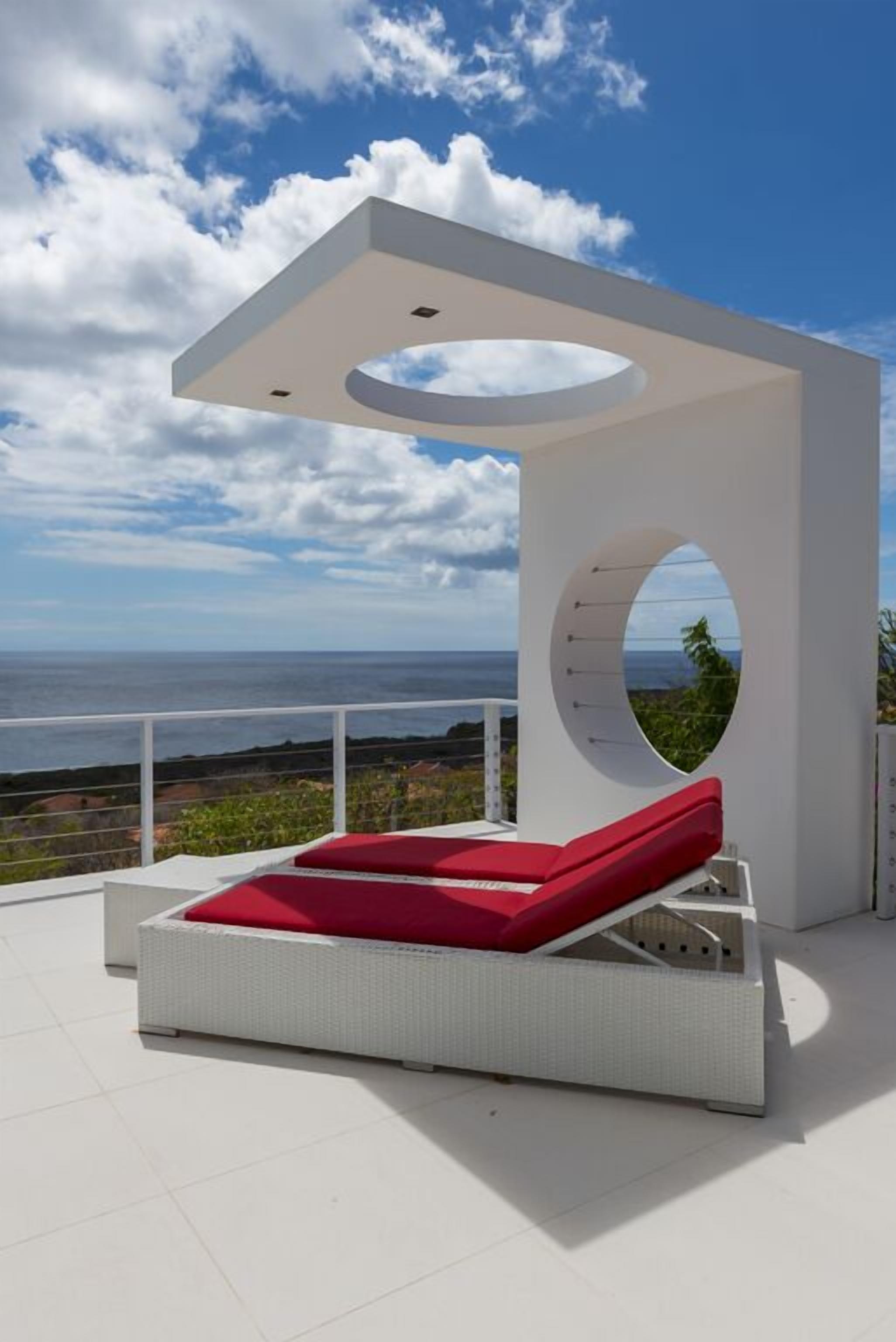 Un espace de détente extérieur moderne, avec un banc rouge doté de coussins, une fenêtre circulaire et une vue sur l’océan.
