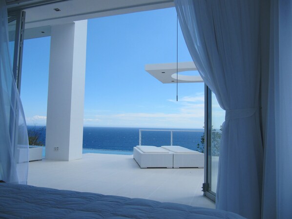 A modern bedroom with a large window overlooking a pool and the sea.