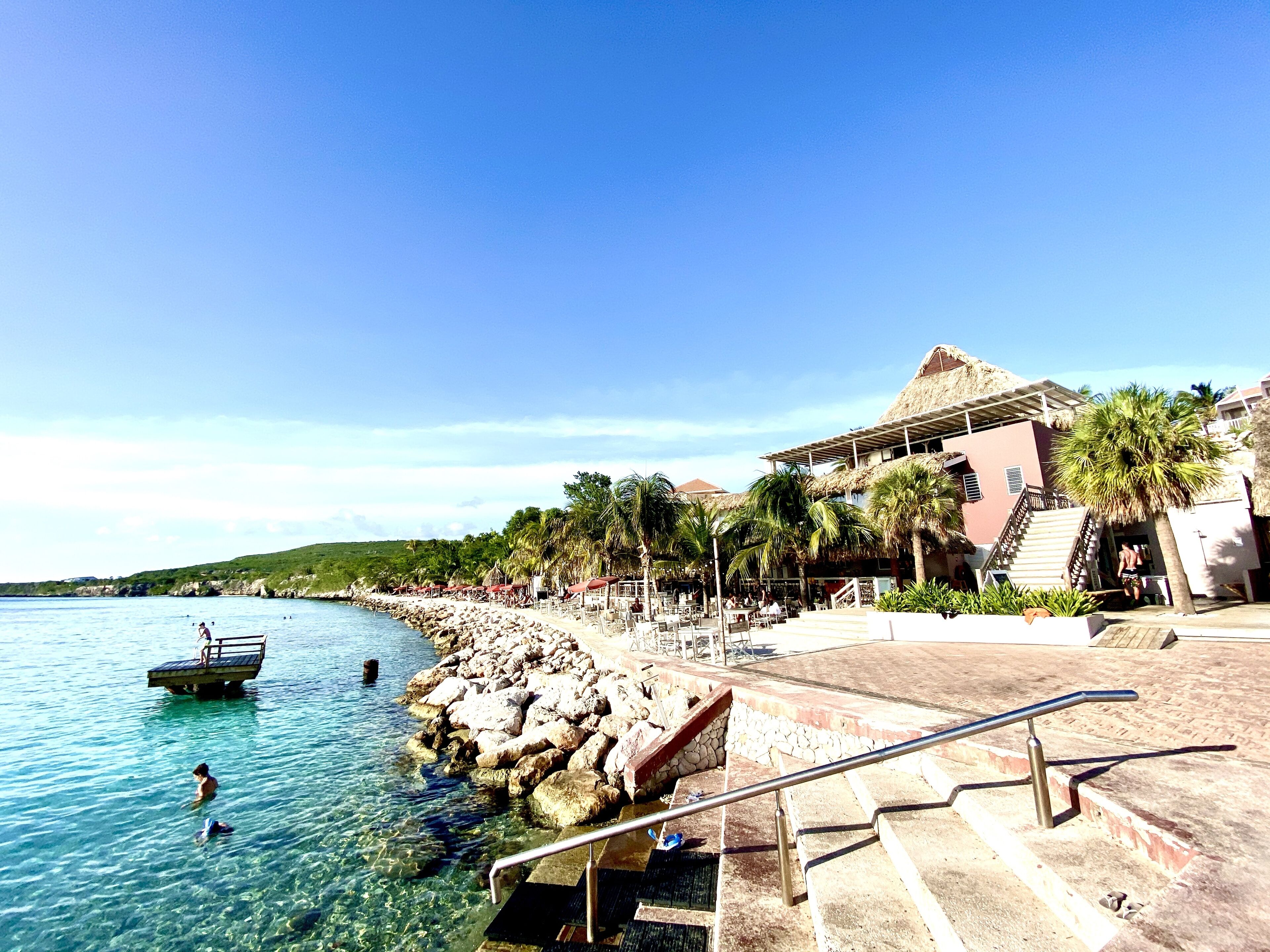 A resort with a clear pool, a beach area, and a staircase leading to a building with a thatched roof.