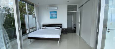 A modern bedroom with a large bed, a glass door leading to a balcony, and a view of greenery outside.