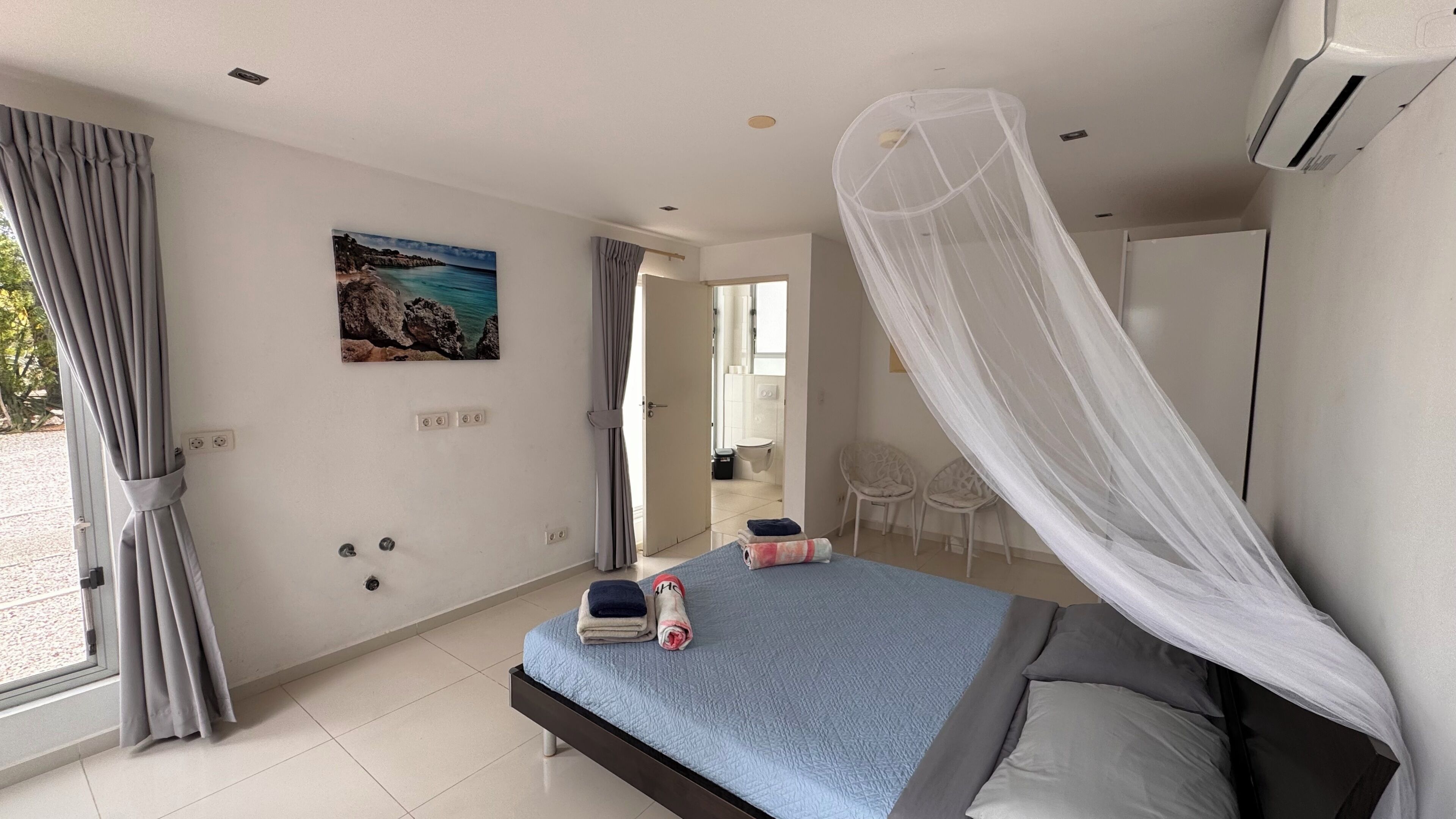 Ein Schlafzimmer mit einem Bett, einem Gemälde, einem Fenster mit Vorhängen und einer Tür zu einem Badezimmer.