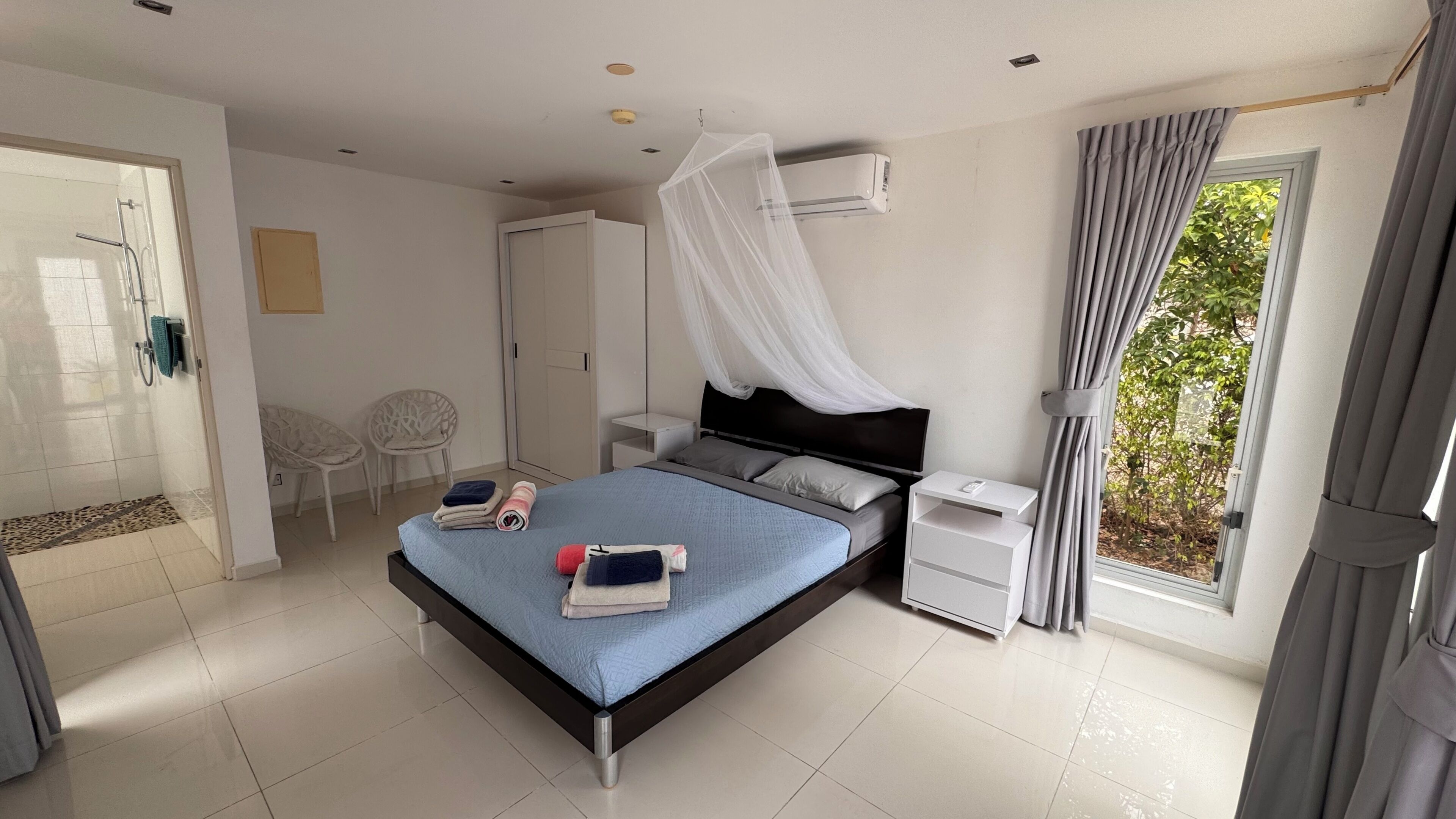 A modern bedroom with a bed, bedside tables, a wardrobe, a shower area, and a view of greenery outside.
