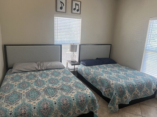 A room with two beds, each with patterned bedding, a nightstand with a lamp, and a window with blinds.