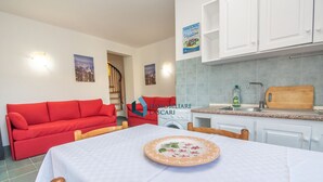 Un espace de vie compact comprenant un canapé rouge, une table à manger avec une assiette à motifs floraux, une cuisine avec un évier et une planche à découper en bois, ainsi qu’une machine à laver.