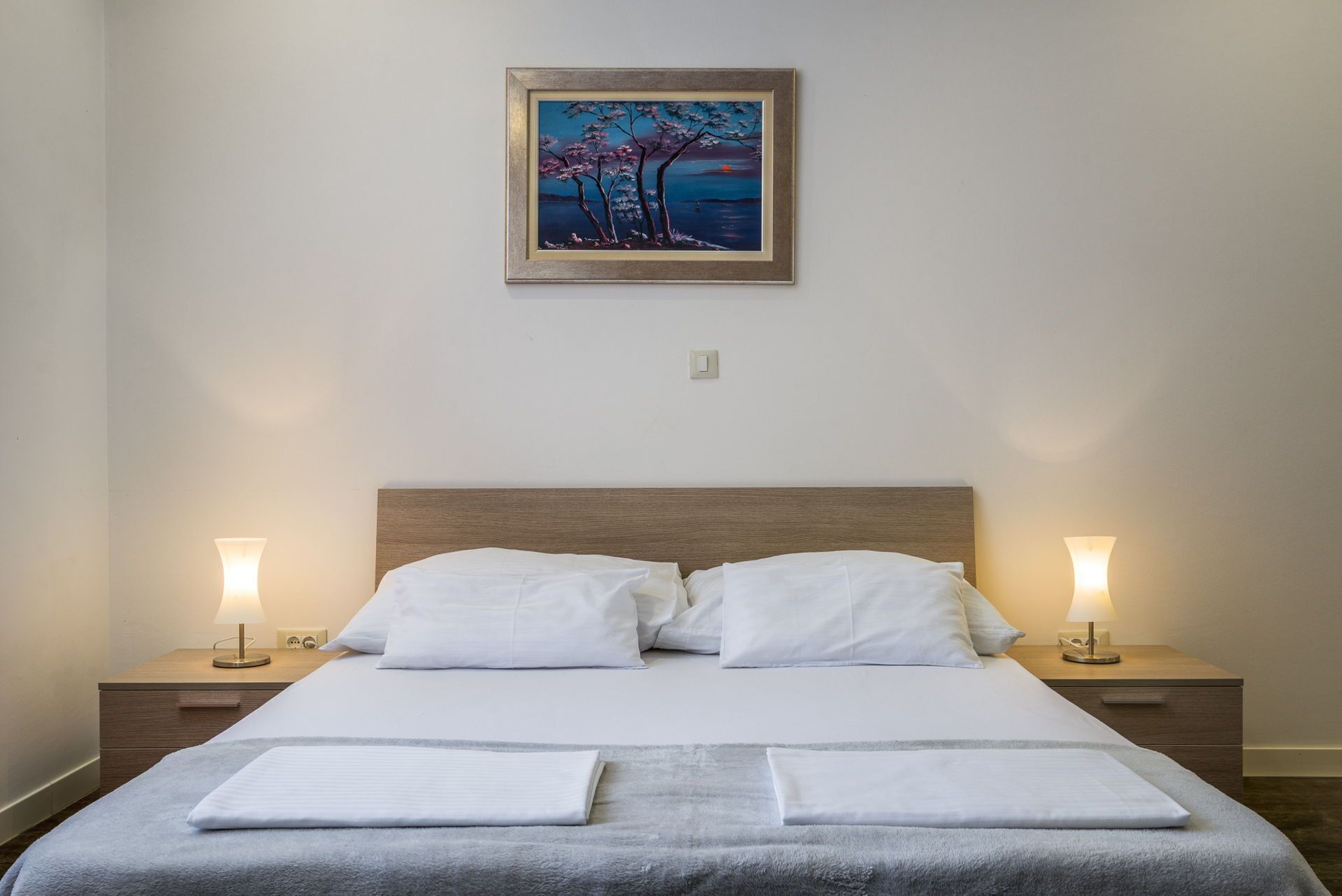 A neatly made bed with white linens and two pillows, flanked by two bedside tables with lamps, in a room with a painting above the bed.