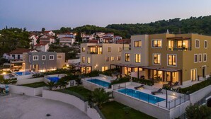A modern multi-story residential building with a swimming pool, outdoor seating, and a view of a hillside.