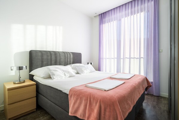 Un dormitorio moderno con cabecera gris, ropa de cama blanca, una mesita de noche de madera con lámpara, y un ventanal con cortinas moradas.