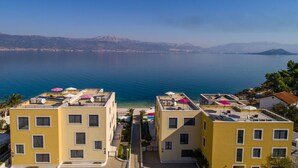 A coastal hotel with multiple yellow buildings, rooftop terraces with umbrellas, and a view of the sea and distant mountains. - Beachfront Apartment Lemonade VI (Trogir)