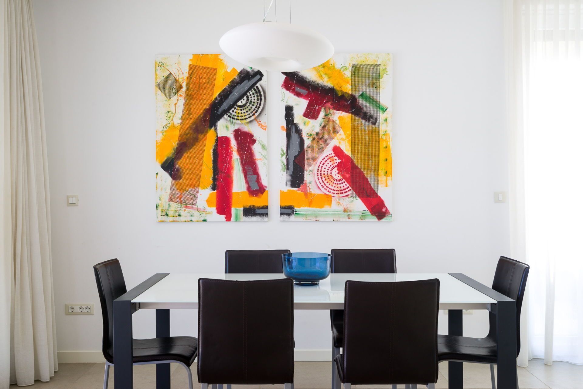 A dining area with a white table and black chairs, a blue bowl on the table, and two abstract paintings on the wall.