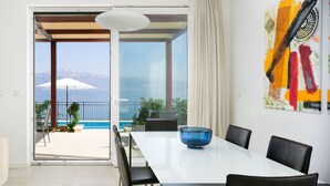 Ein stilvoller Essbereich mit Blick auf das Meer und die Berge, ein großes abstraktes Gemälde an der Wand und eine blaue Glasvase auf dem Tisch.