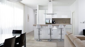 Eine moderne Küche mit Einbauofen, Kaffeemaschine und einem Edelstahlkühlschrank. Es gibt drei Barhocker und einen Esstisch mit Stühlen.
