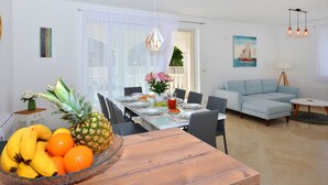 Ein Essbereich mit Holztisch, Stühlen und einer Obstschale. Im Wohnbereich befindet sich eine Couch und eine Lampe.