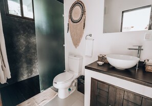 A bathroom with a white toilet, a beige macramé wall hanging, a white sink with a vessel basin, and a dark wooden vanity. - Xcacel Dreams Jungle Suites & Spa (Tulum)