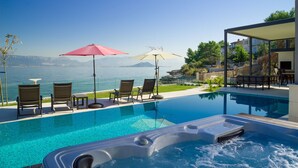 Une piscine à débordement avec un jacuzzi, des chaises longues et des parasols, offrant une vue sur un lac et des montagnes au loin.