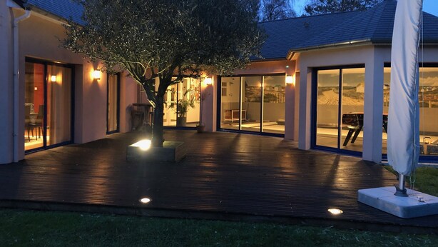 A modern house with a wooden deck, large glass doors, and a tree in the center.