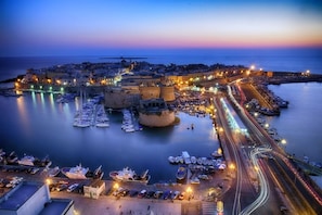 A coastal cityscape at dusk with a prominent fortress, marina, and busy roads.