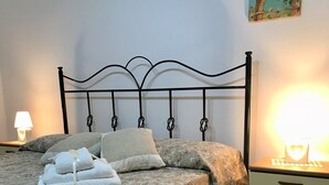 A neatly made bed with a floral bedspread, a headboard with a unique design, a nightstand with a lamp, and a wall painting. - Apartment On the Beaches of Gallipoli (Gallipoli)
