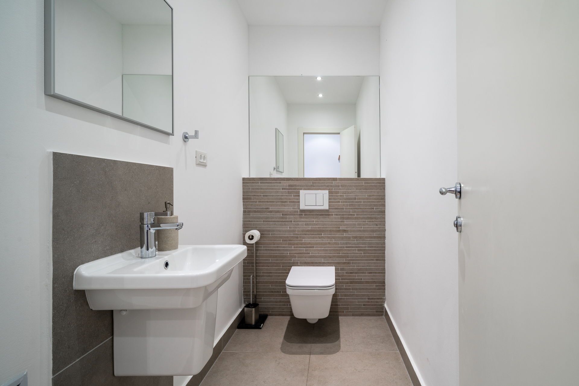 Ein schmales Badezimmer mit einem weißen Waschbecken, einer Toilette und einem Spiegel an der linken Wand.