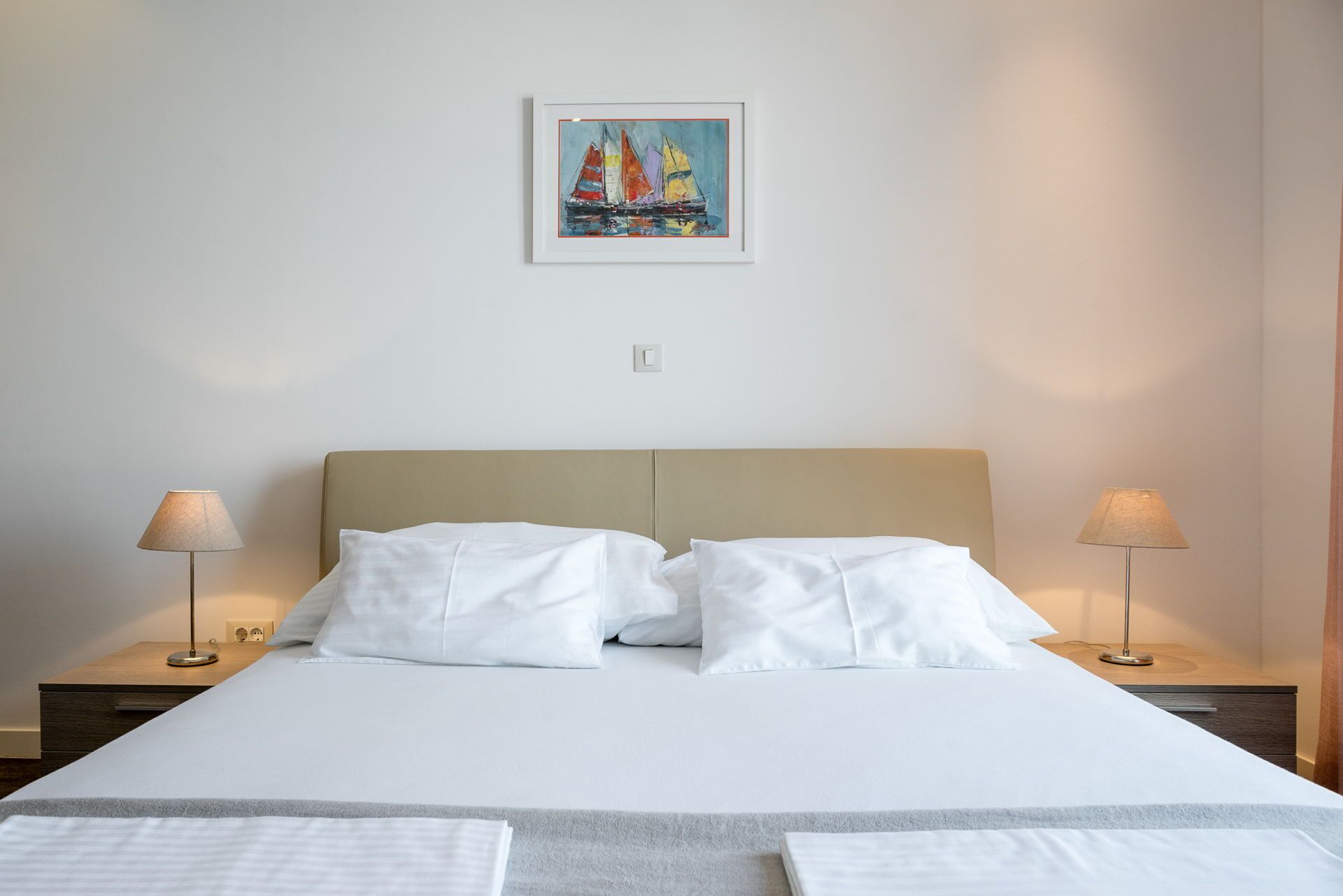 Un letto rifatto con lenzuola bianche, due lampade sui comodini e un quadro appeso al muro raffigurante dei velieri.
