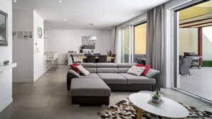 Ein modernes Wohnzimmer mit einer grauen Eckcouch, einem kleinen runden Tisch und einer großen Schiebetür, die zu einem Außenbereich führt.