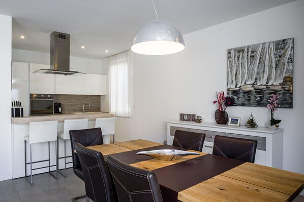 Cocina moderna con armarios blancos, horno empotrado y zona de comedor con mesa de madera y sillas de cuero negro.