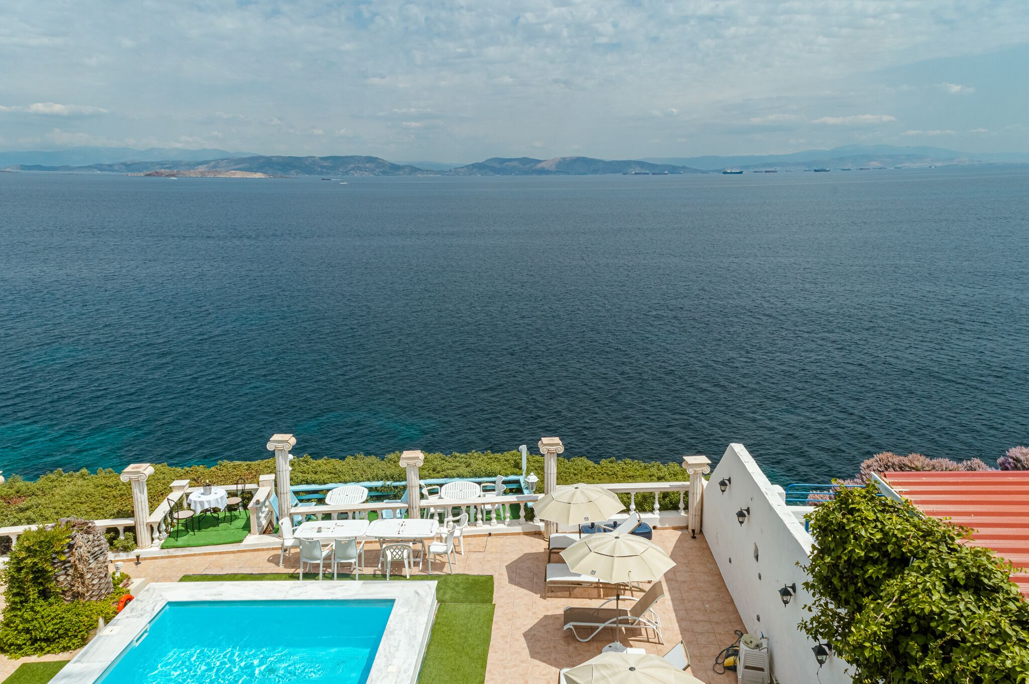 Zona de piscina con vistas al mar, mobiliario exterior y un edificio blanco con columnas.