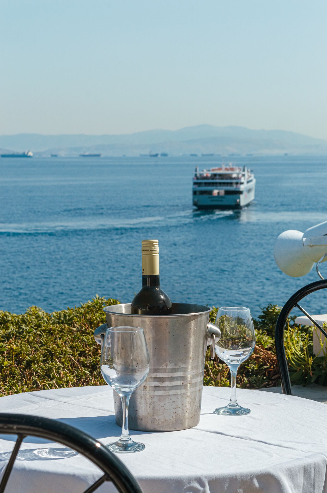 Une table avec une bouteille de vin et des verres, donnant sur une étendue d’eau où l’on aperçoit un navire au loin.