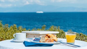 Ein Tisch mit einer weißen Tasse, einem Teller mit Croissants und Kuchen, einem Glas Orangensaft und einer Serviette, vor einer Kulisse aus Meer und Grünpflanzen.