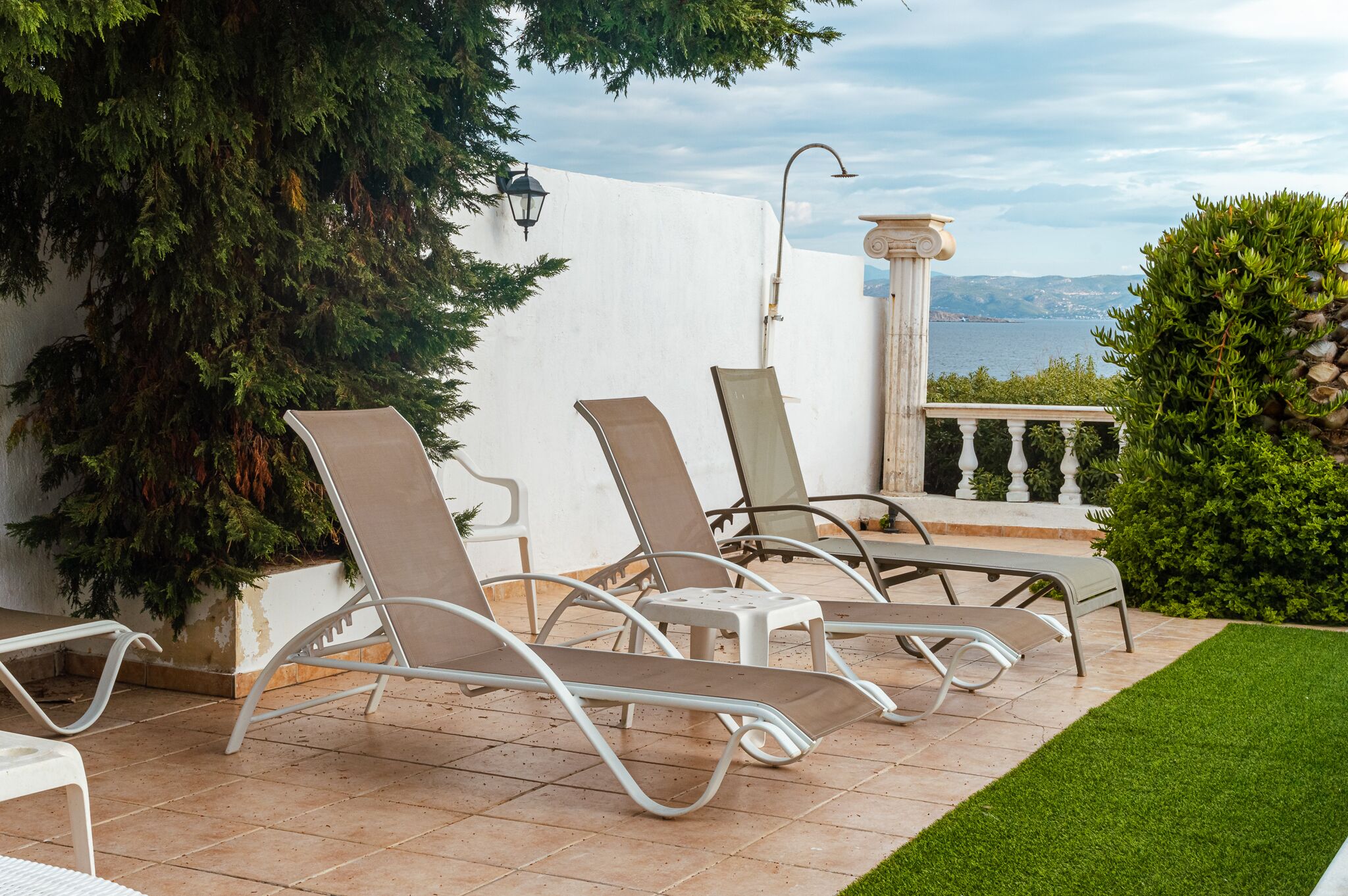 Une terrasse avec trois fauteuils de jardin, un mur blanc, un arbre en pot et une vue sur la mer.