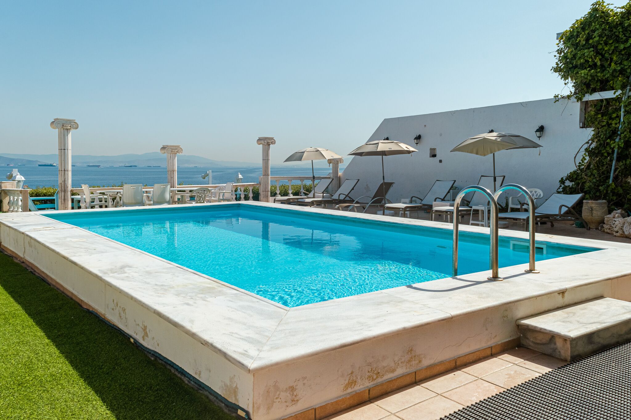 A rectangular swimming pool with clear blue water, surrounded by white columns and lounge chairs with umbrellas.