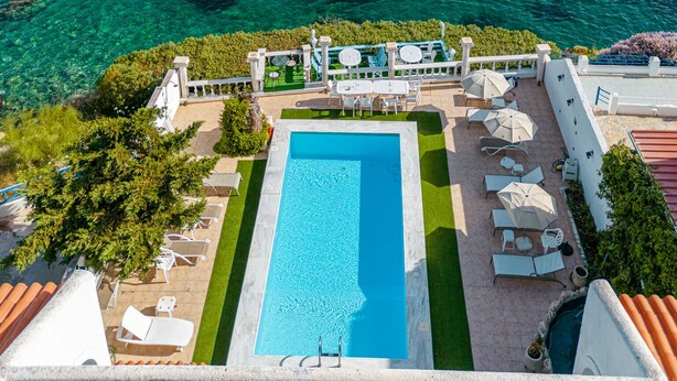 Ein rechteckiger Pool mit klarem, blauem Wasser, umgeben von einer Terrasse mit weißen Liegestühlen und Sonnenschirmen, mit Blick auf eine weite Wasserfläche und entfernte Hügel.