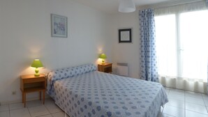 A bedroom with a bed, two bedside tables with lamps, a radiator, a window with curtains, and a framed picture on the wall.