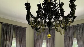 Una habitación con una gran lámpara de araña, un piano, una mesa con un florero y un sillón estampado.