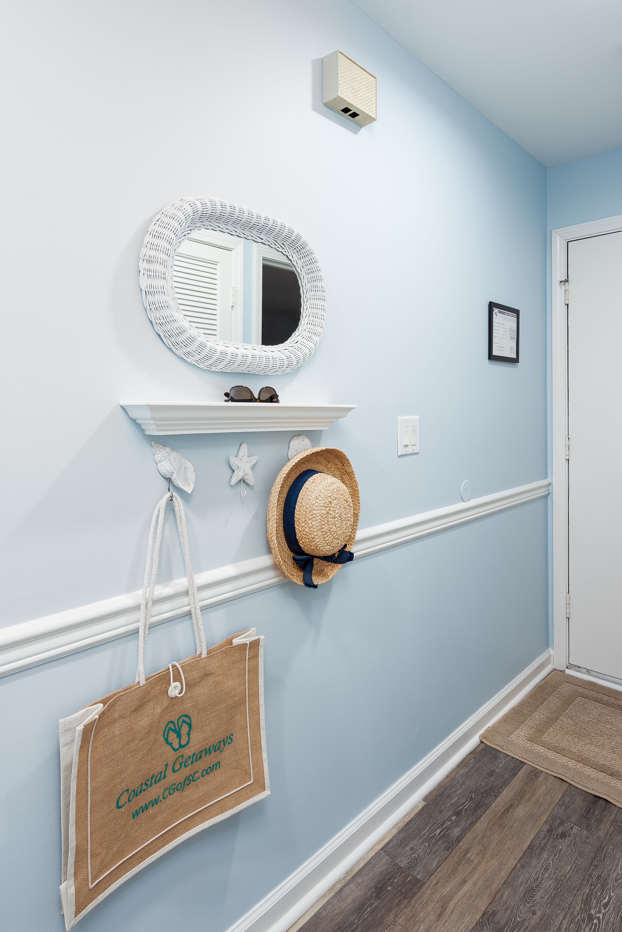 A hallway with a light blue wall, a white-framed mirror, a shelf with sunglasses, a straw hat, and a beach-themed tote bag.