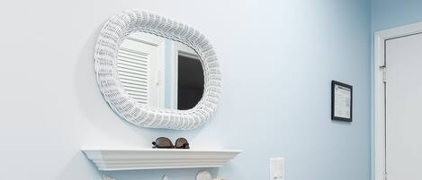 A hallway with a light blue wall, a white-framed mirror, a shelf with sunglasses, a straw hat, and a beach-themed tote bag.