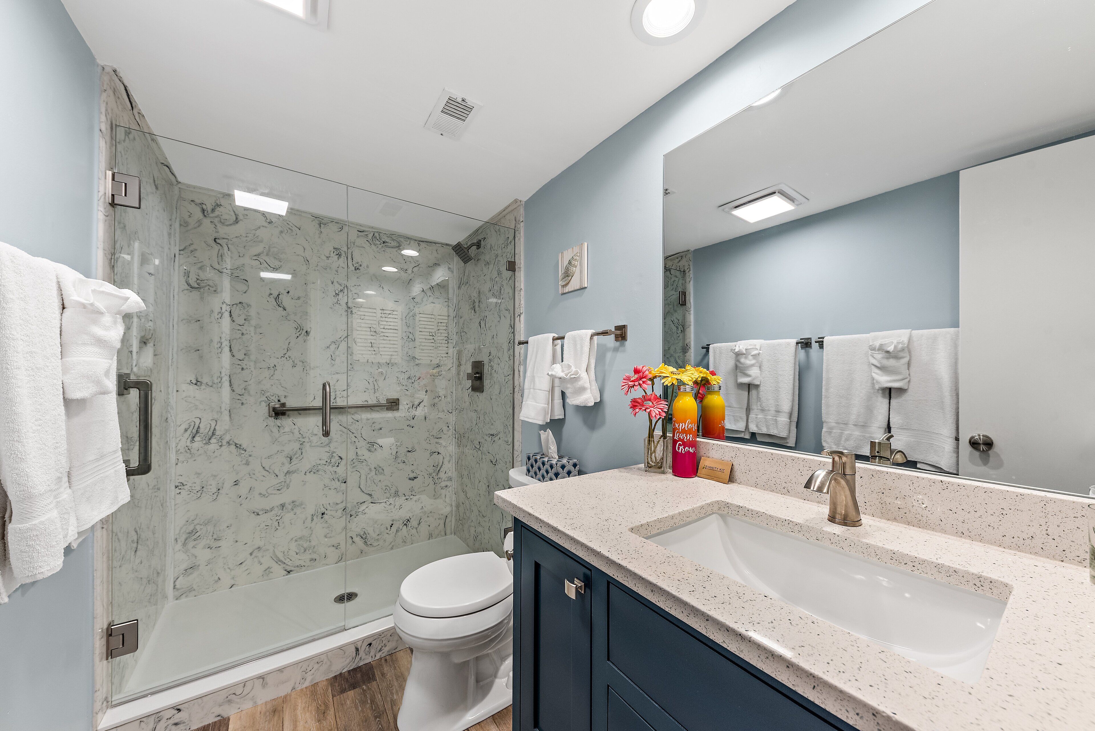 Une salle de bain moderne avec une douche à parois de verre, des toilettes blanches, un lavabo doté d’un plan de travail en marbre et un grand miroir.