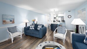 Sala de estar com um sofá azul, poltronas brancas e uma mesa de centro. Há uma cozinha ao fundo.