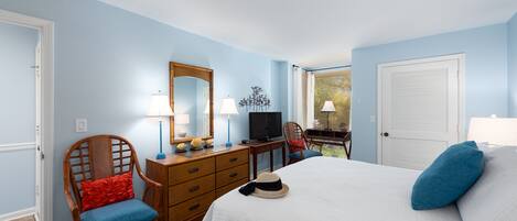 Quarto com cama, cadeira de madeira, cômoda, televisão e janela com persianas.