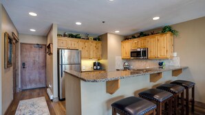A kitchen with wooden cabinets, a stainless steel refrigerator, a microwave, and a granite countertop. There are three bar stools and a small plant on the countertop.