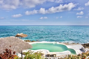 A pool with a clear blue edge, surrounded by a white deck and a thatched roof structure.
