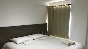 En snyggt bäddad säng med vita sängkläder och en hoprullad handduk, en mörk trägav och ett fönster med beige gardiner.