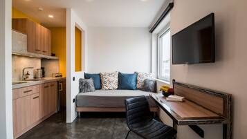 A compact room with a kitchenette, a bed with colorful pillows, a black chair, and a mounted TV.