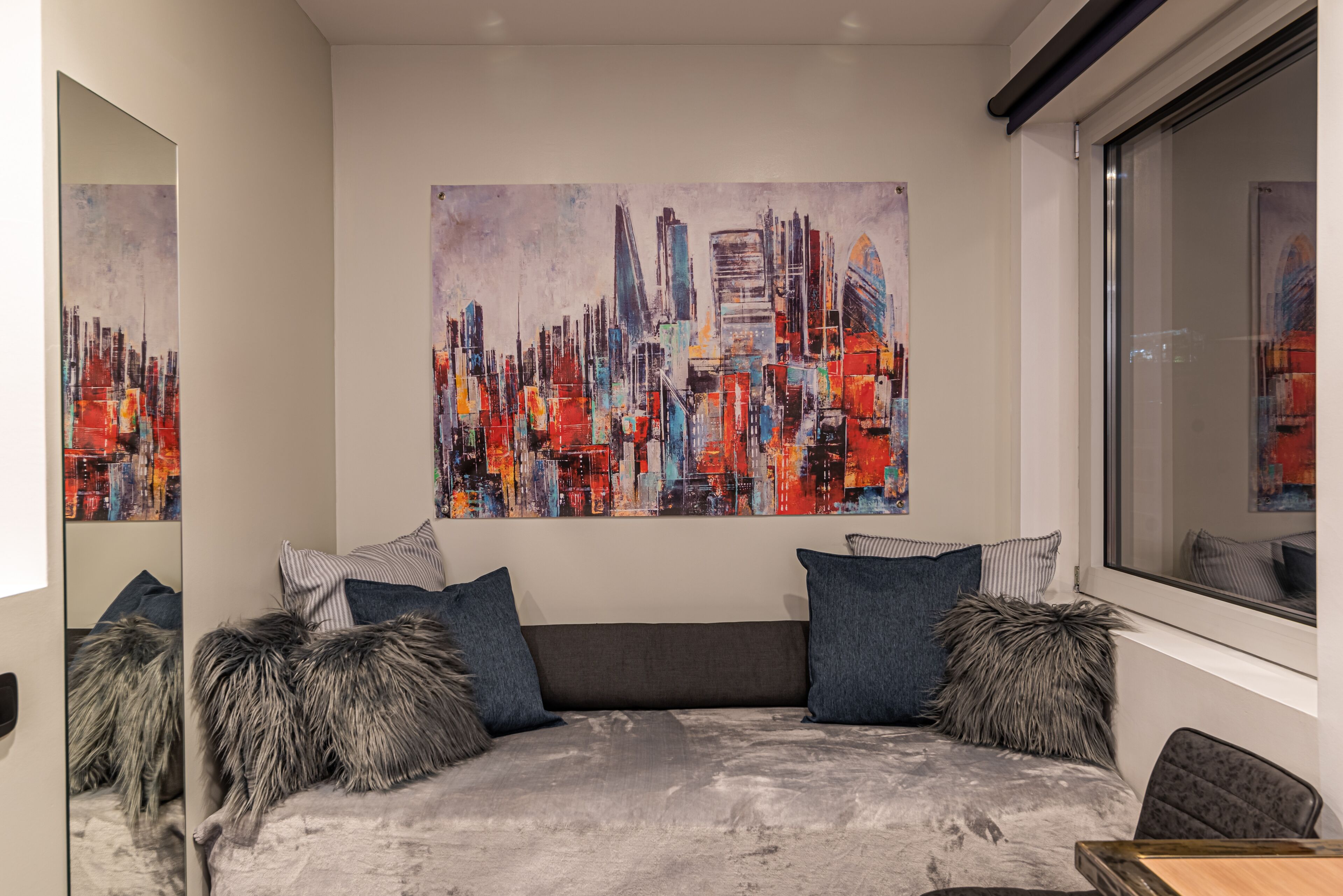 A modern living room with a large cityscape painting, a gray sofa with patterned cushions, and a window with a view.