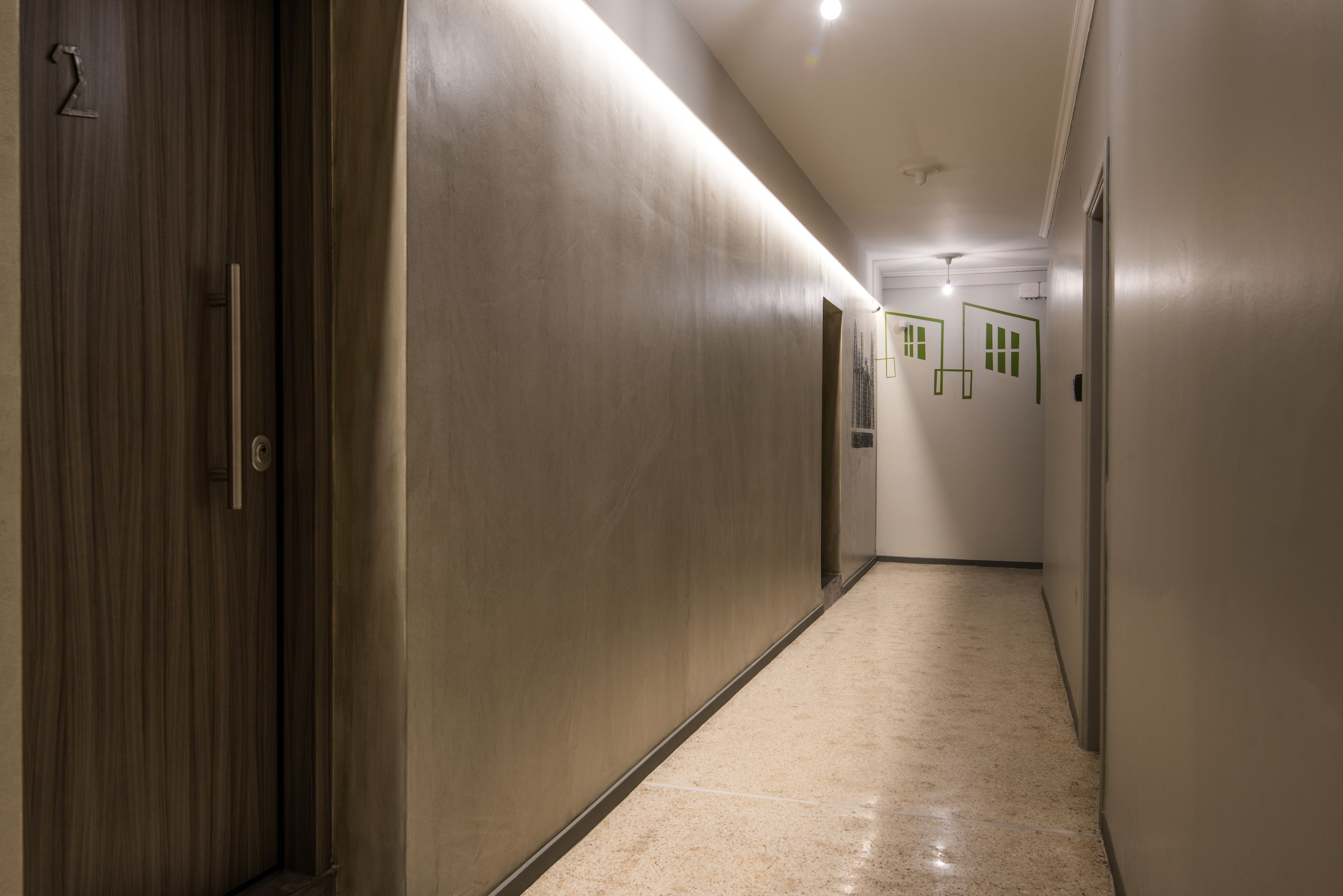 A long, narrow hallway with a wooden door on the left and a series of doors on the right. The floor is tiled, and the walls are painted in a light color.