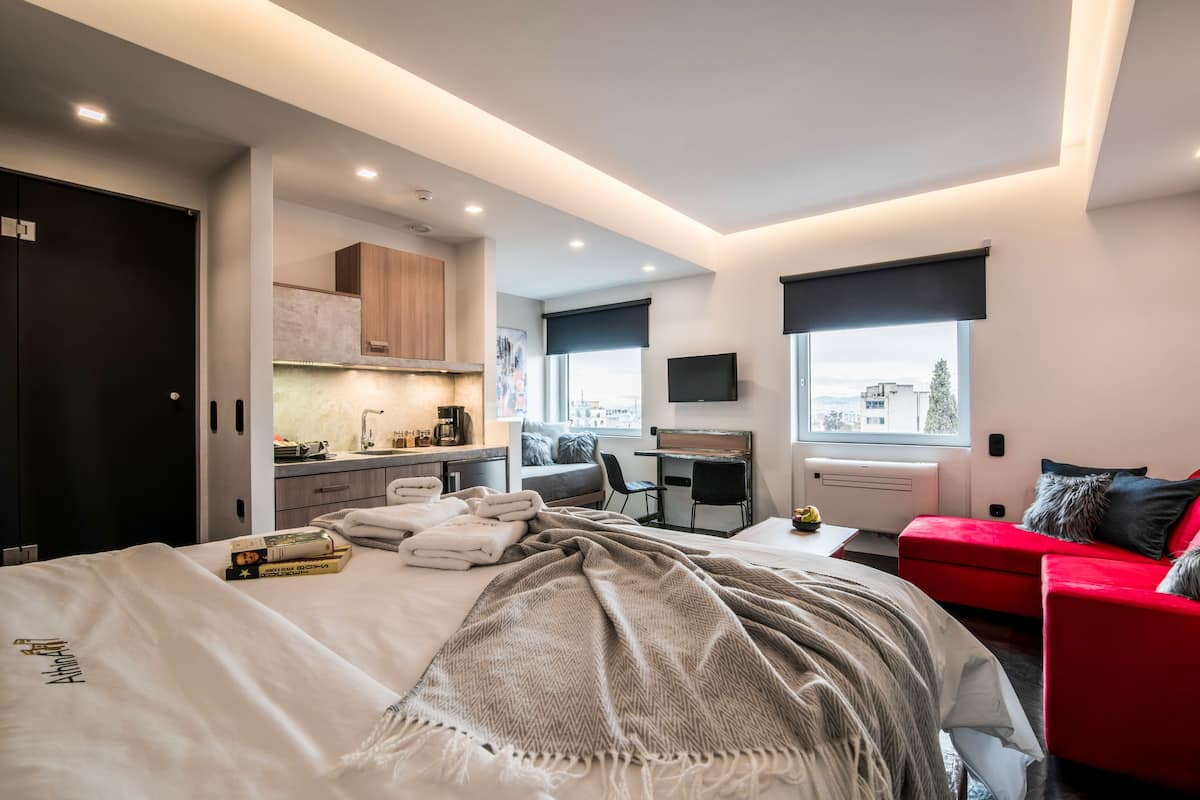 A modern bedroom with a bed, a desk, a red sofa, and a view of the city.